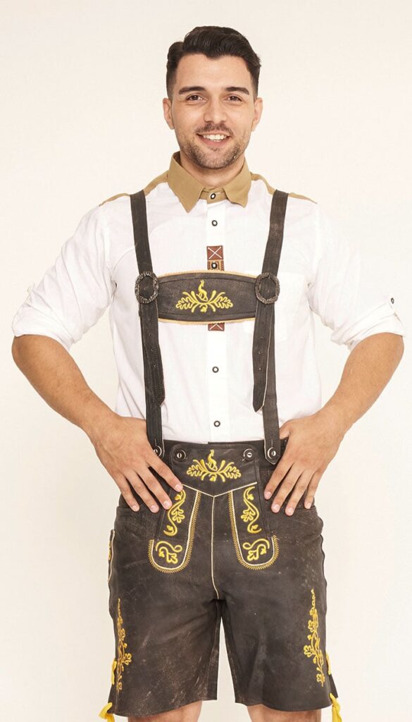 Front view of a man standing with hands on hips wearing dark brown Lederhosen with vibrant yellow embroidery and a white shirt with a tan collar. - Lederhosen For Sale