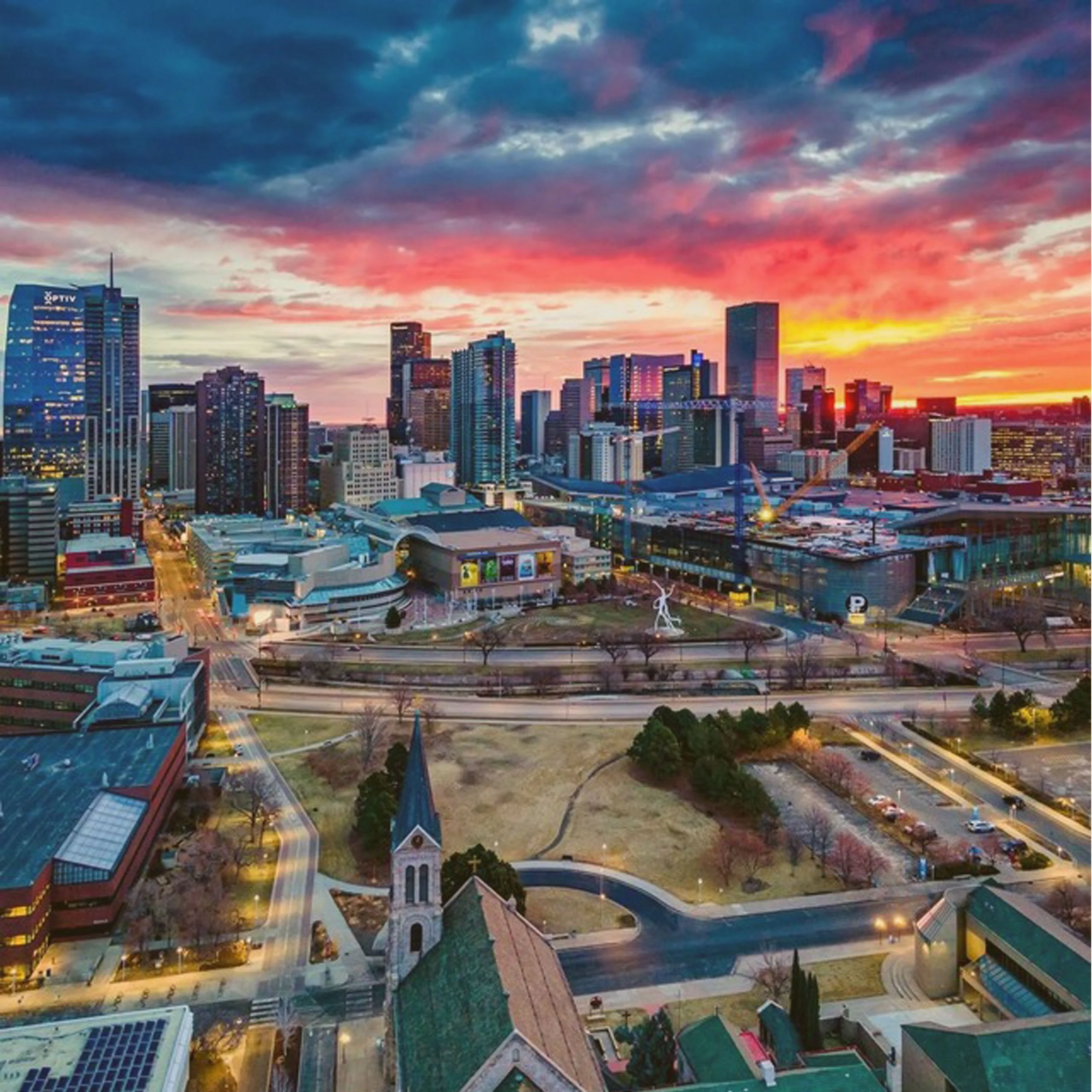 High-angle aerial view of the Denver, Colorado city skyline featuring modern skyscrapers under a vibrant orange and pink sunset sky. - Lederhosen For Sale