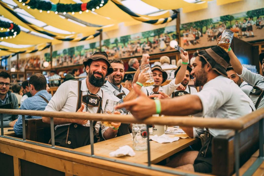 Men in traditional Lederhosen celebrating with beer mugs inside an Oktoberfest tent. - Lederhosen For Sale