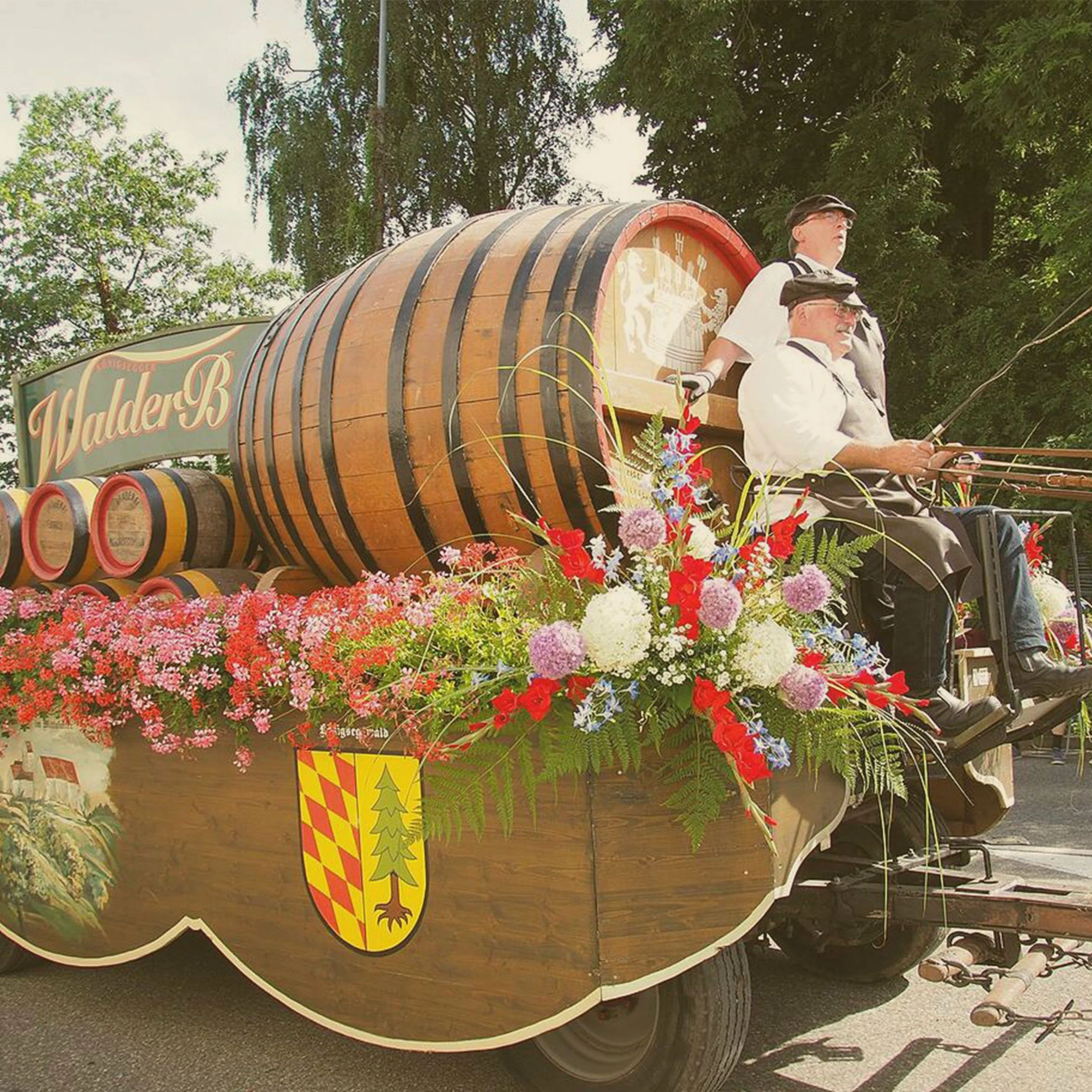 A decorated festival float with large wooden beer barrels, floral arrangements, and men steering it during an Oktoberfest parade. - Lederhosen For Sale