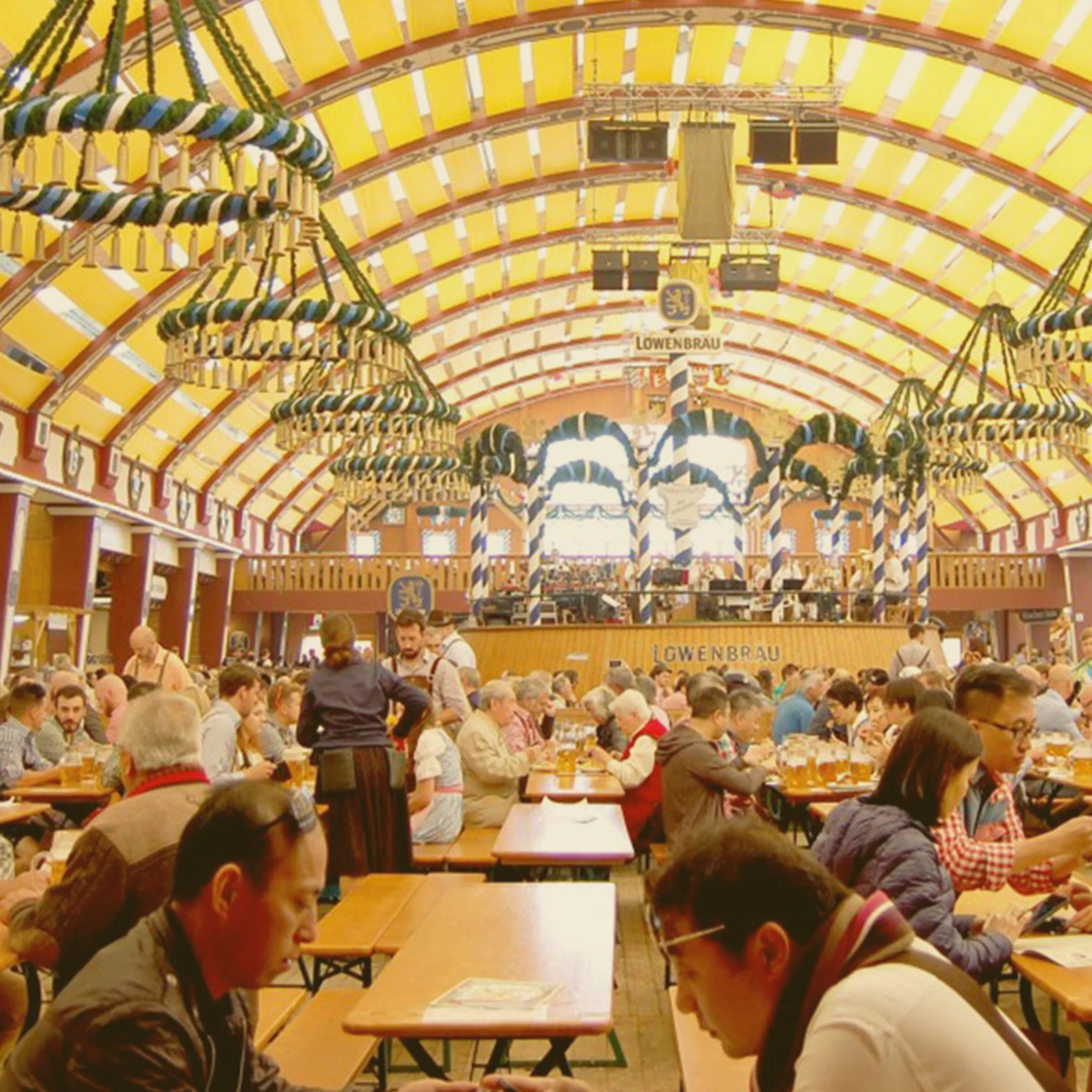 A crowded interior of a Löwenbräu beer tent filled with people dining at long tables during an Oktoberfest celebration in Kansas. - Lederhosen For Sale