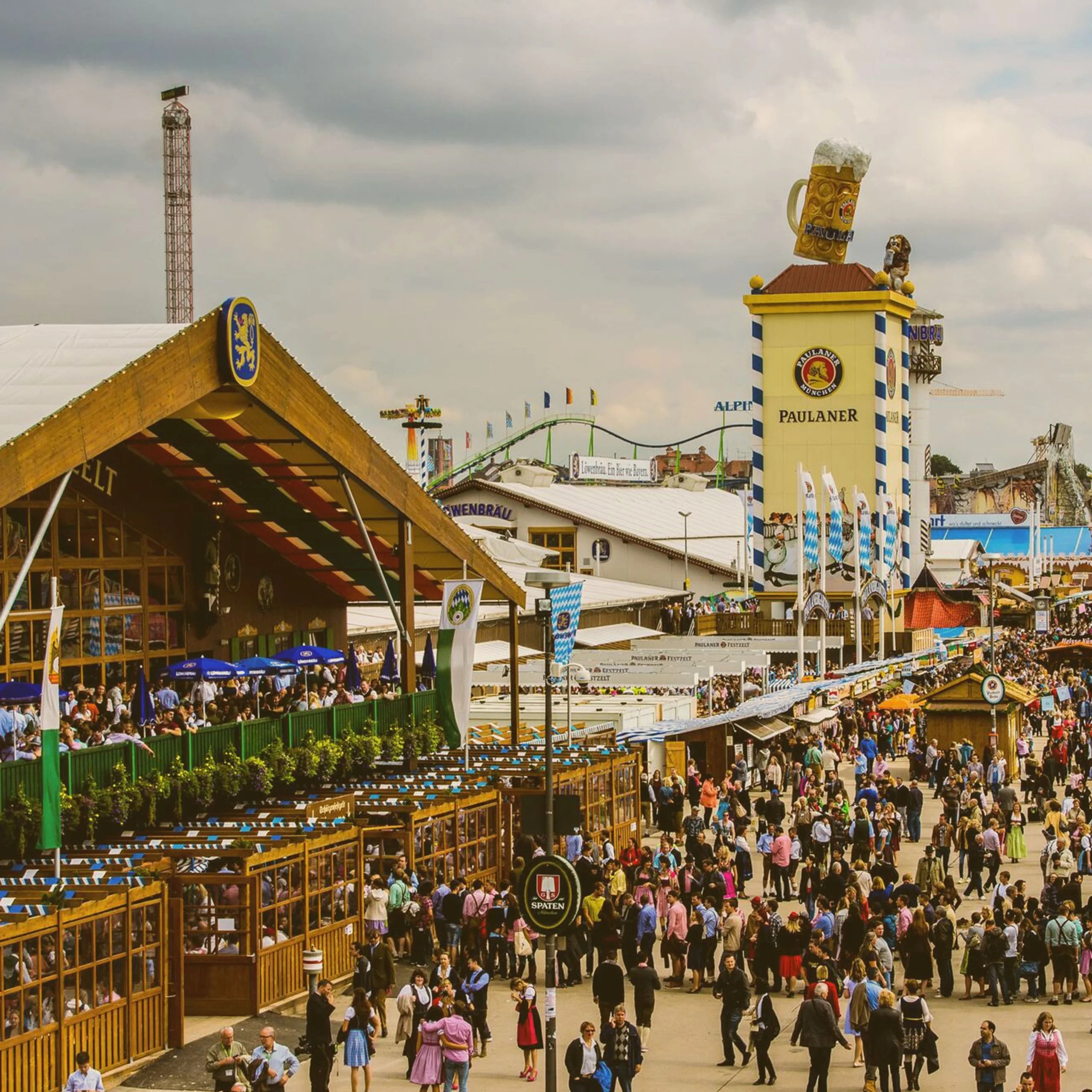 Crowded outdoor festival grounds at Oktoberfest featuring a large Paulaner beer tower and traditional wooden beer tents under a cloudy sky. - Lederhosen For Sale