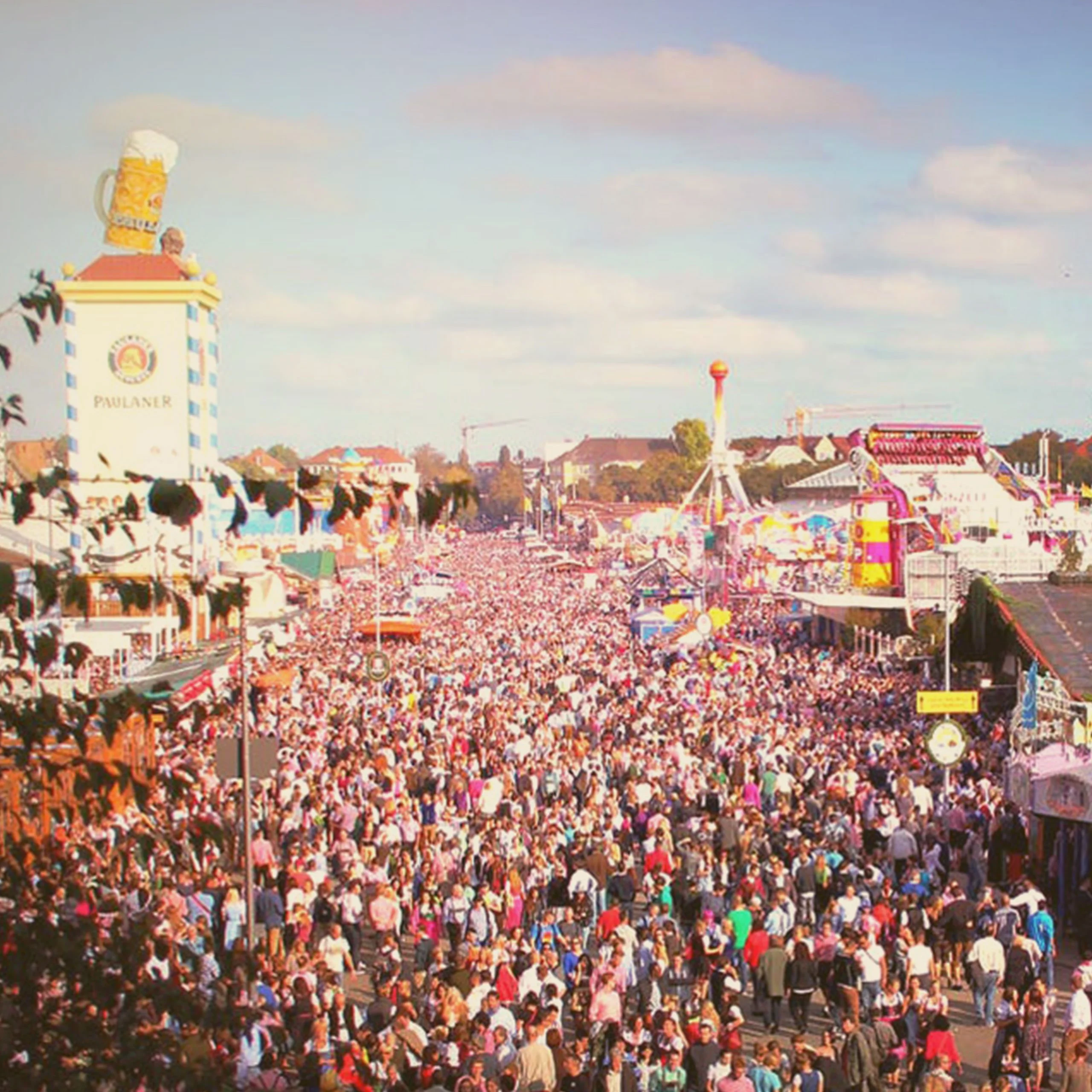 A massive crowd of people filling the festival grounds at an Oktoberfest event in Mississippi, featuring the Paulaner beer tower and amusement park rides. - Lederhosen For Sale