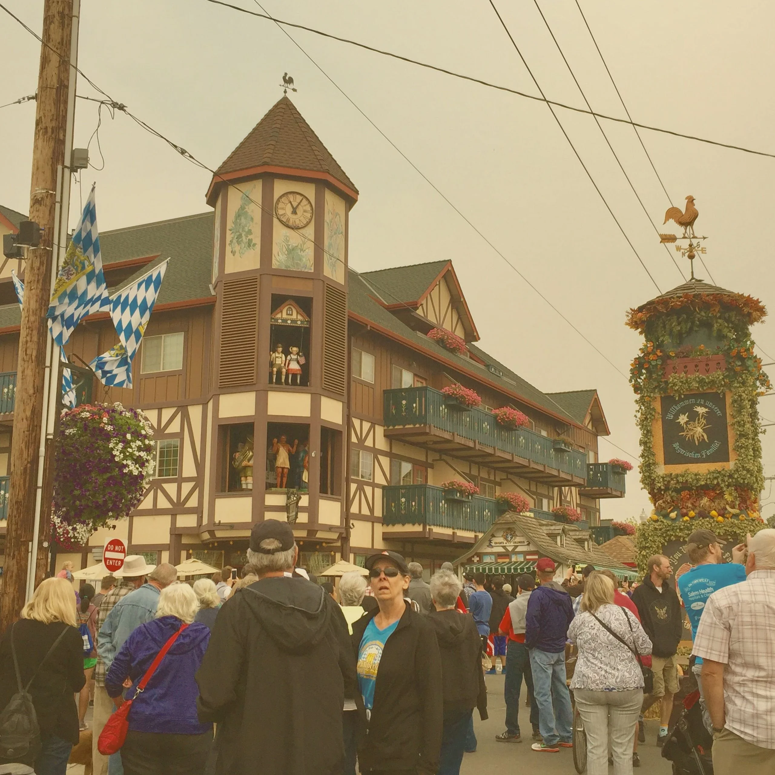 A large crowd gathered in front of traditional Bavarian-style buildings and a glockenspiel tower under a hazy sky during the Mt. Angel Oktoberfest. - Lederhosen For Sale