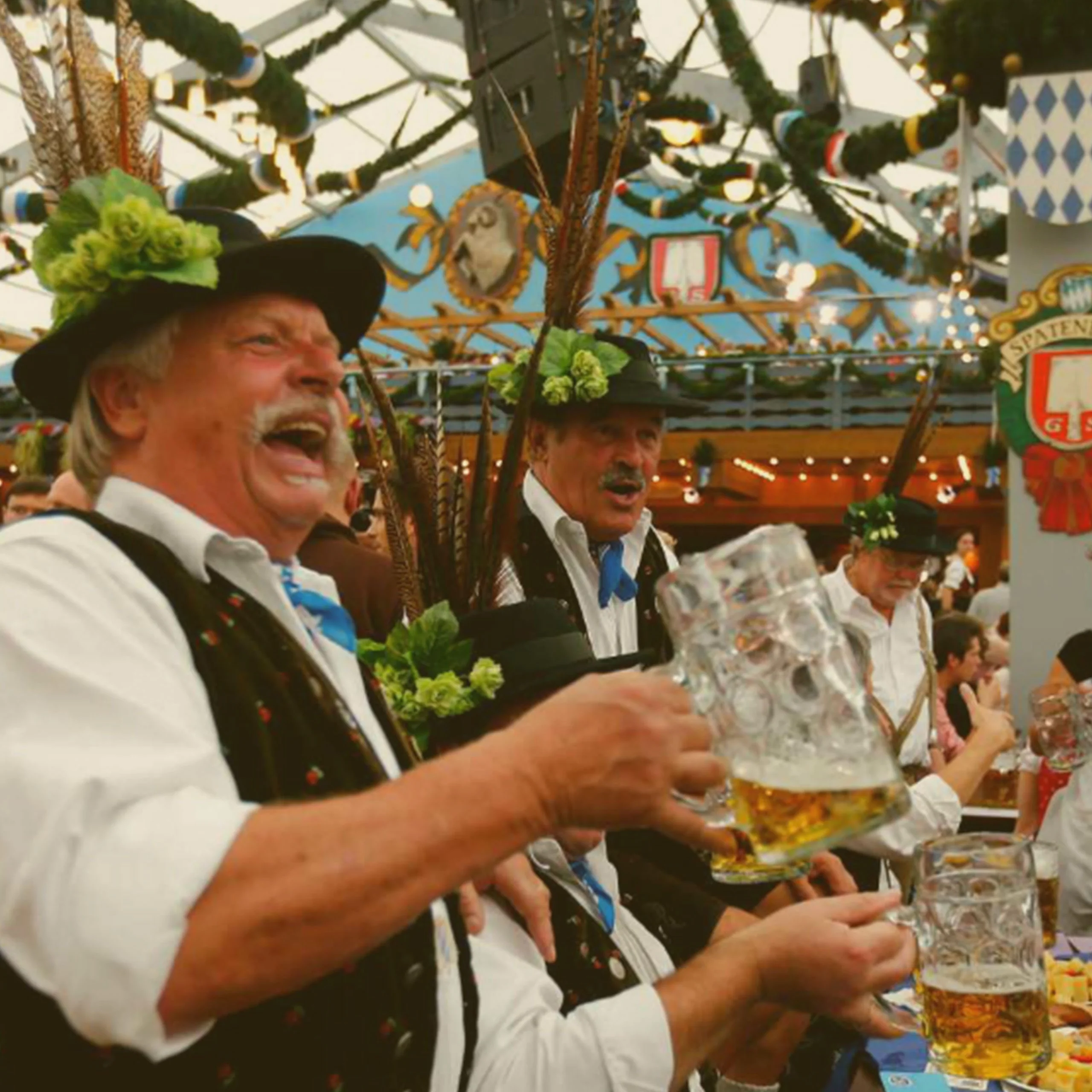 Men in traditional Bavarian hats with feathers singing and holding beer mugs inside a crowded Spaten festival tent at Oktoberfest in Munich, Germany. - Lederhosen For Sale