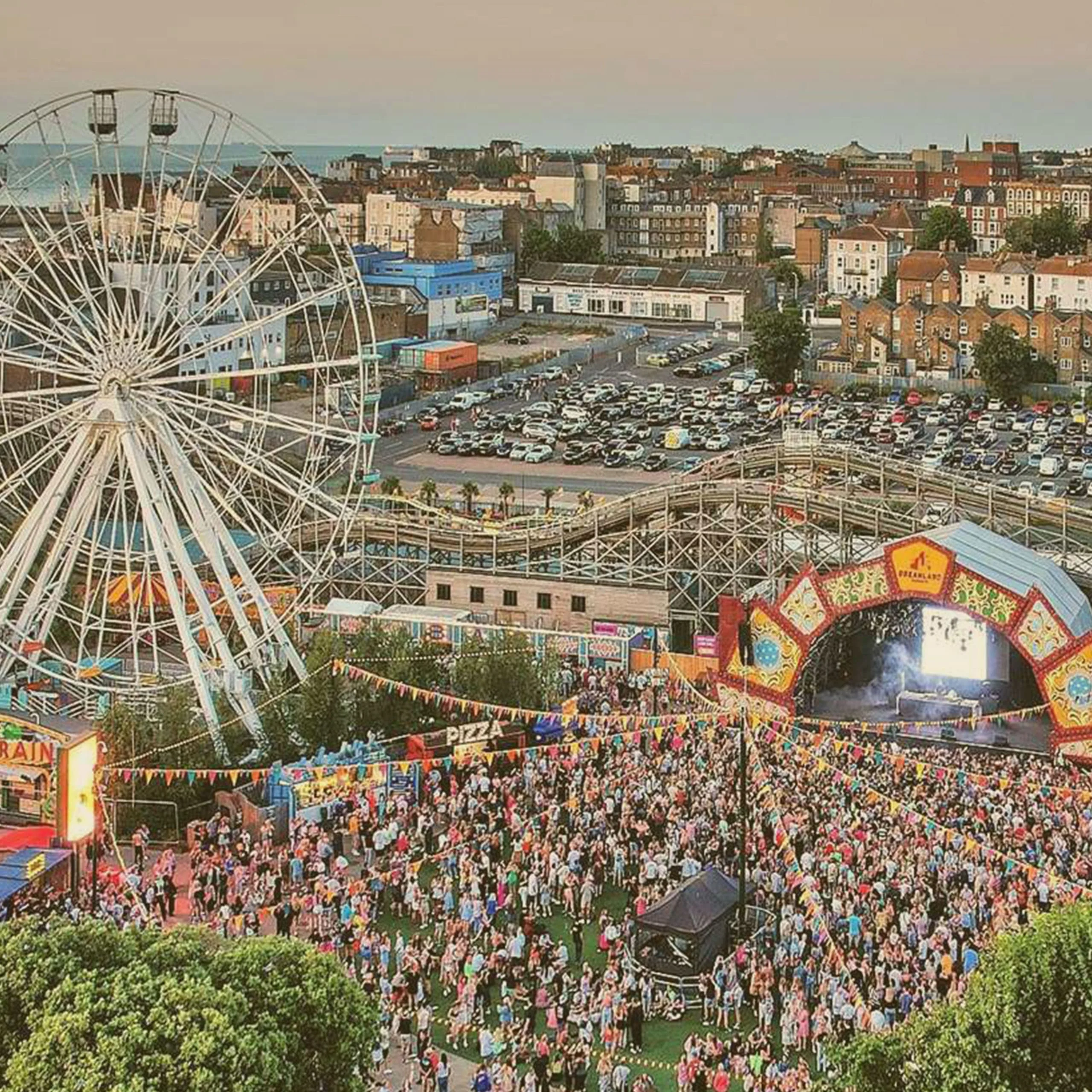 High-angle view of a crowded outdoor music festival at Dreamland Margate amusement park, featuring a giant Ferris wheel, wooden roller coaster, and a large stage. - Lederhosen For Sale