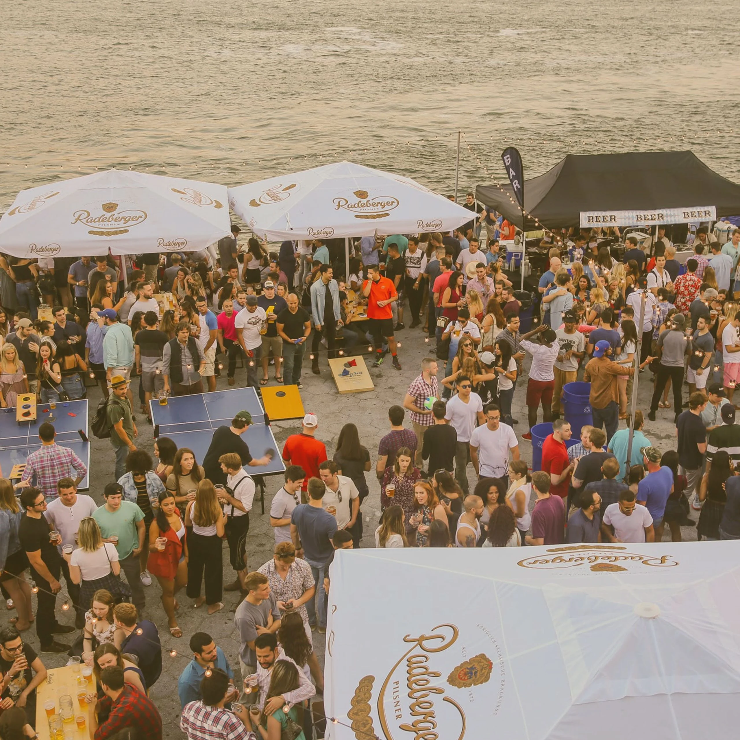 A large outdoor crowd celebrating at a riverside Oktoberfest event in New Mexico, featuring Radeberger beer umbrellas, ping-pong tables, and beer tents. - Lederhosen For Sale