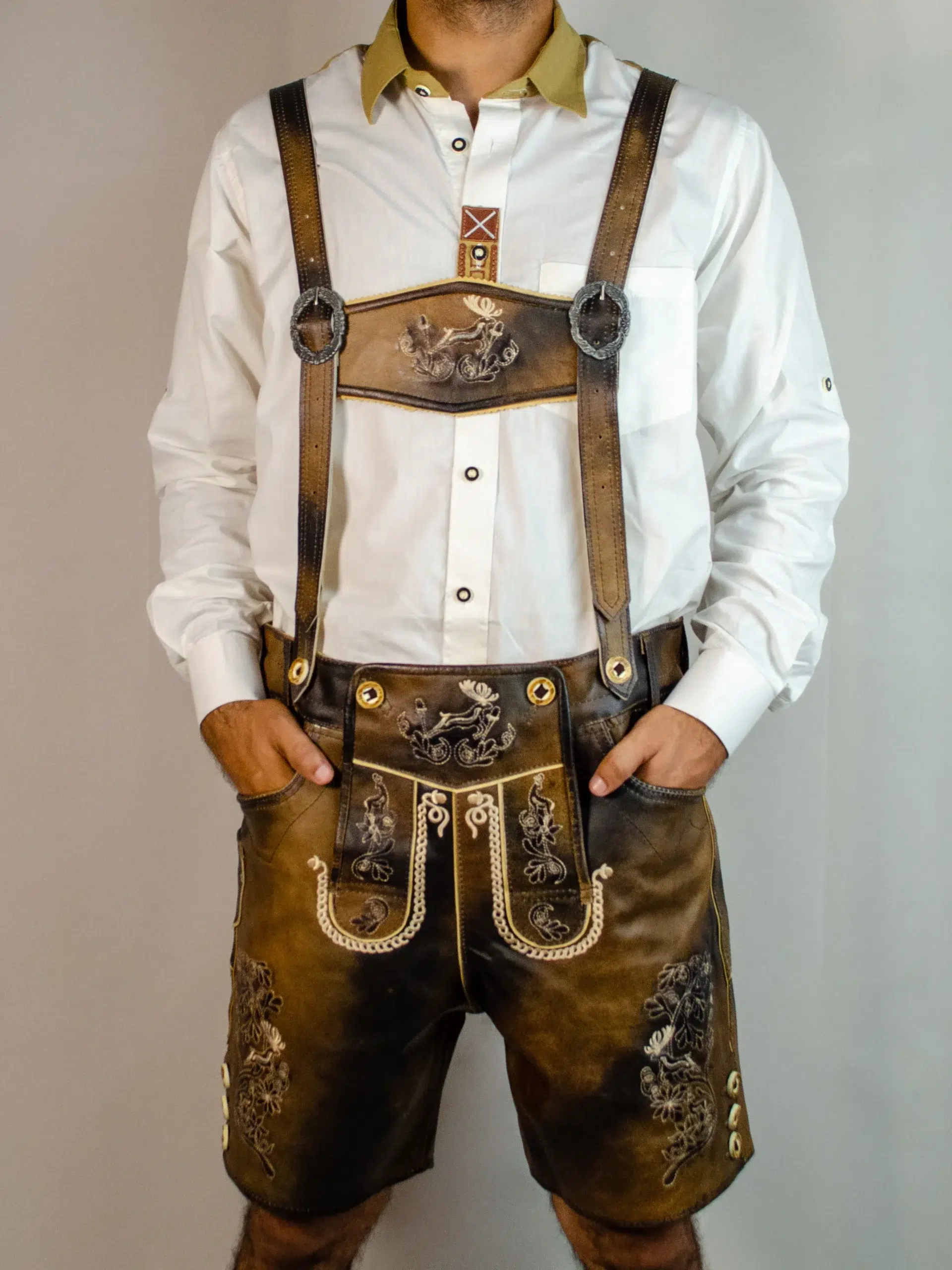 Front view of a man standing with hands in pockets wearing dark chocolate distressed leather Lederhosen with matching stag-embroidered suspenders and a white shirt. - Lederhosen For Sale