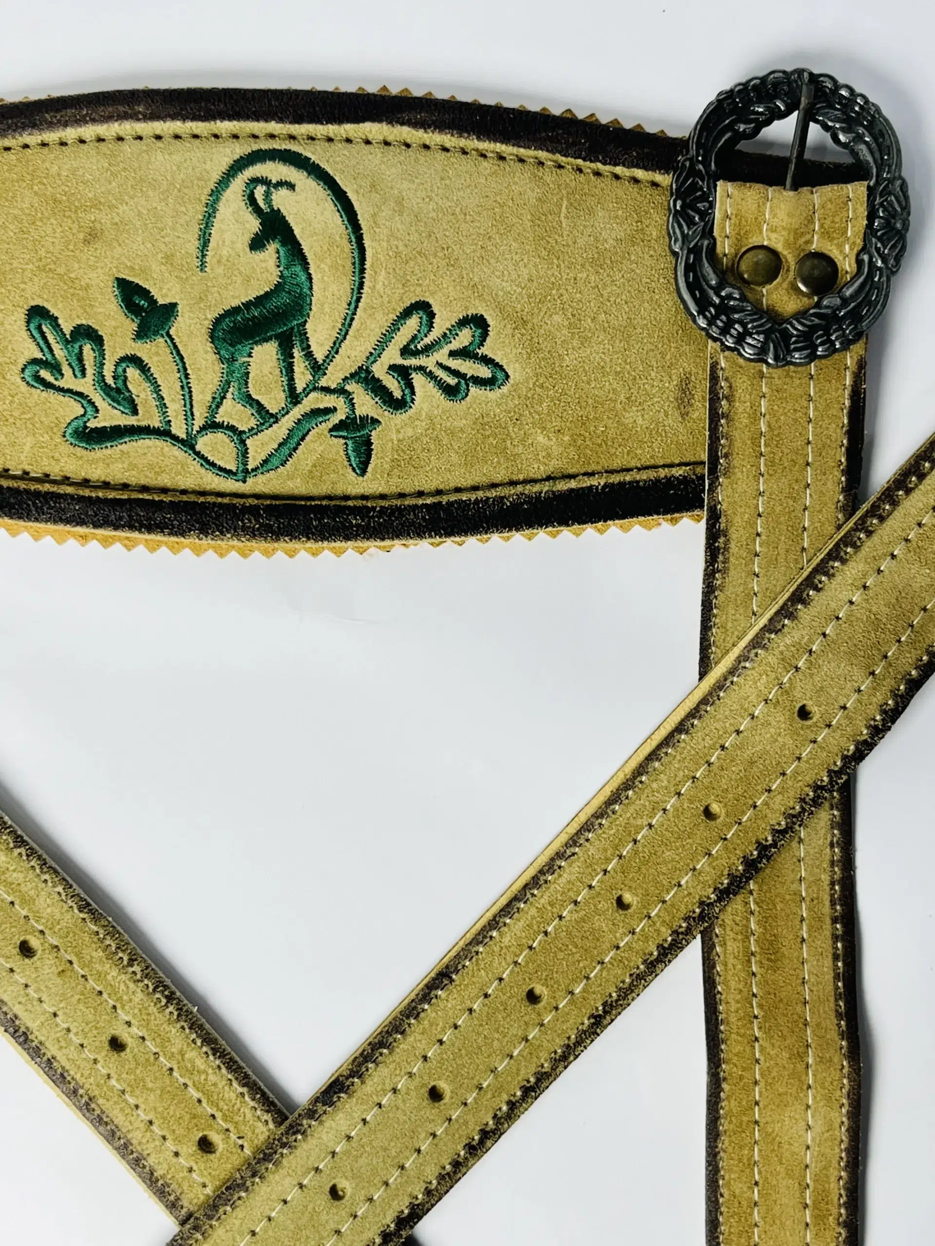 Close-up of green stag and oak leaf embroidery on the front cross-bridge of light gold Lederhosen suede suspenders with a detailed dark silver metal buckle. - Lederhosen For Sale