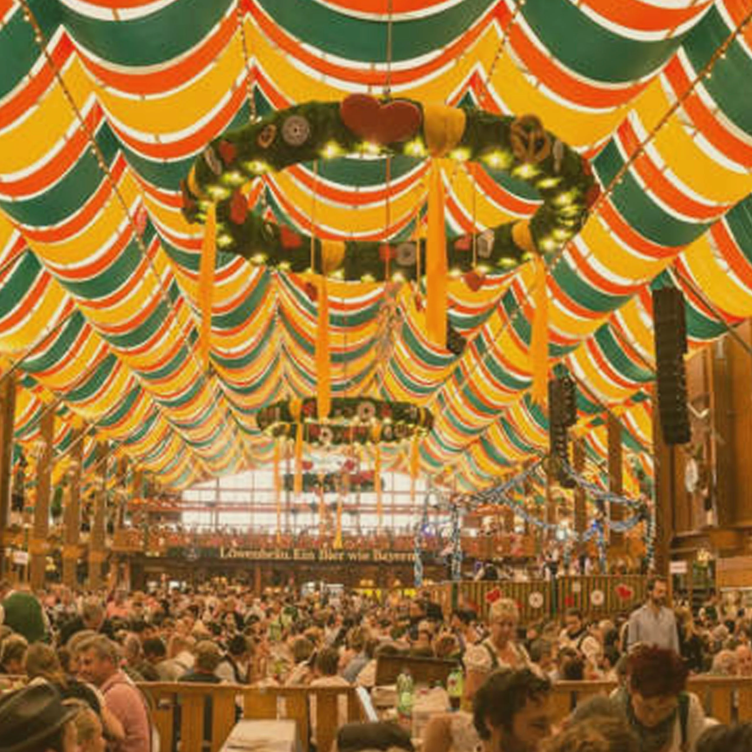 Traditional Oktoberfest beer tent crowded with people, featuring green, orange, and yellow draped fabric ceilings and large decorative wreaths. - Lederhosen For Sale