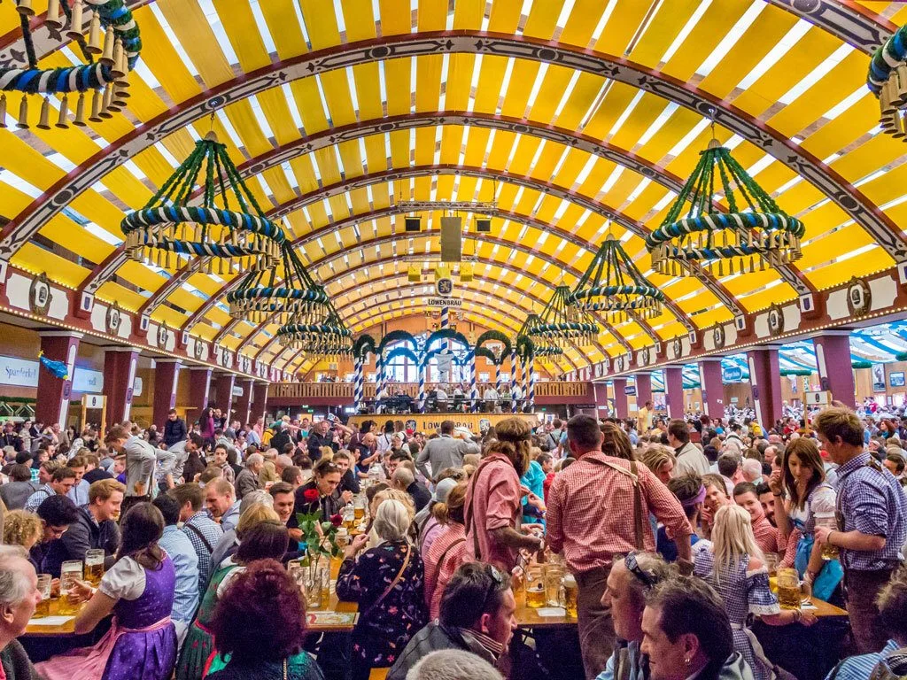 Crowded interior of a massive Oktoberfest beer tent with a yellow striped ceiling, hanging decorations, and people celebrating. - Lederhosen For Sale