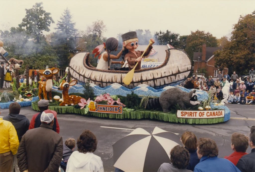 A large festival parade float featuring figures in a canoe, a raccoon, and deer. - Lederhosen For Sale